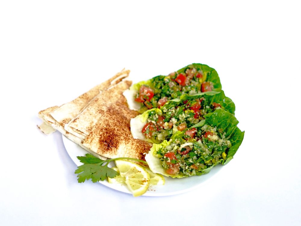 Frontalsicht eines Tellers, worauf sich Tabouleh-Salat auf Salatblätter mit Fladenbrot daneben und zwei halbe Scheiben Zitrone mit einem Blatt Petersilie als Dekoration zu sehen sind.